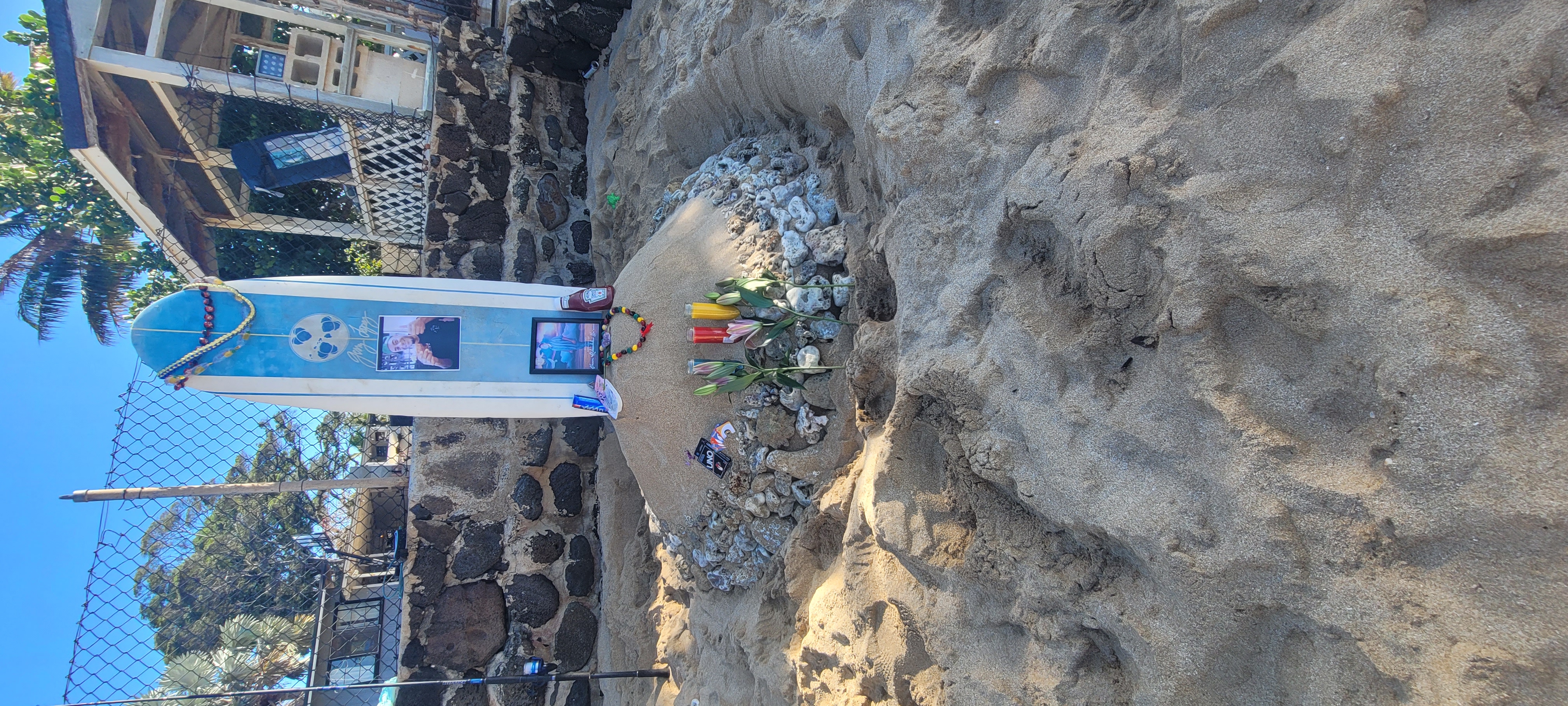 A mound of sand and rocks, with lei and offerings at the base of a surfboard.