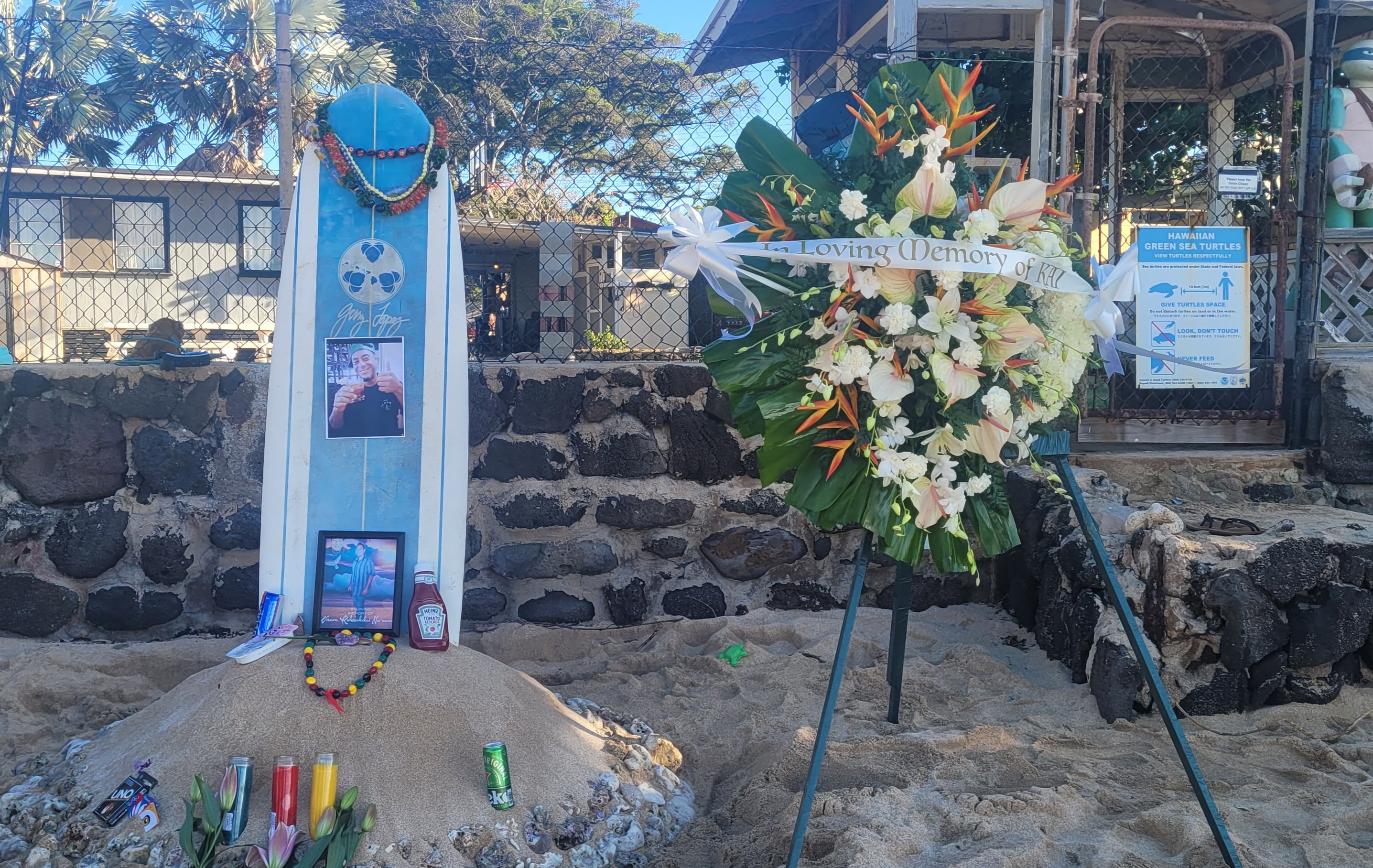A mound of sand and rocks, with lei and offerings at the base of a surfboard. A wreath now stands to the right