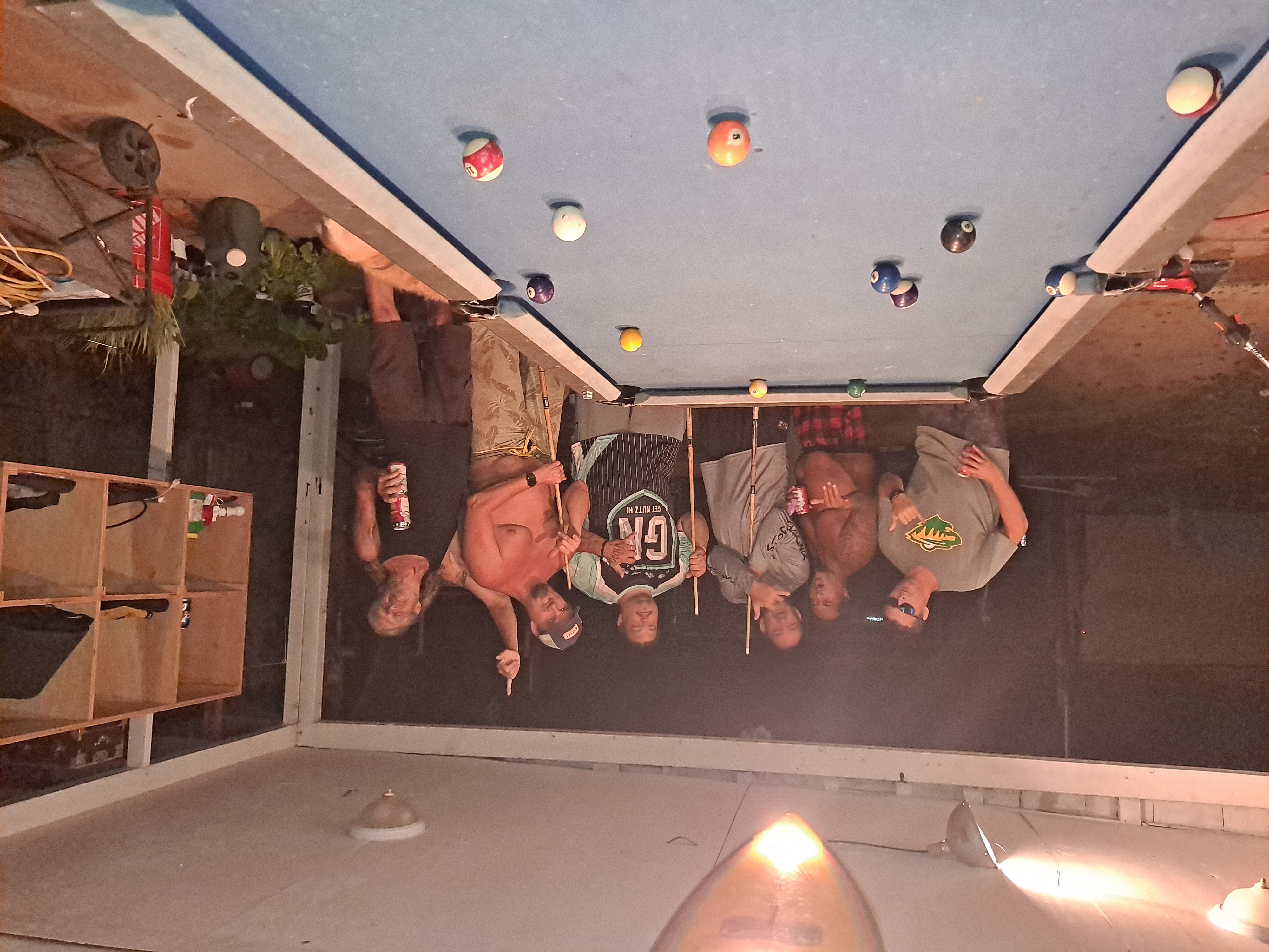 Six men, holding beers and throwing shakas, standing over a pool table.