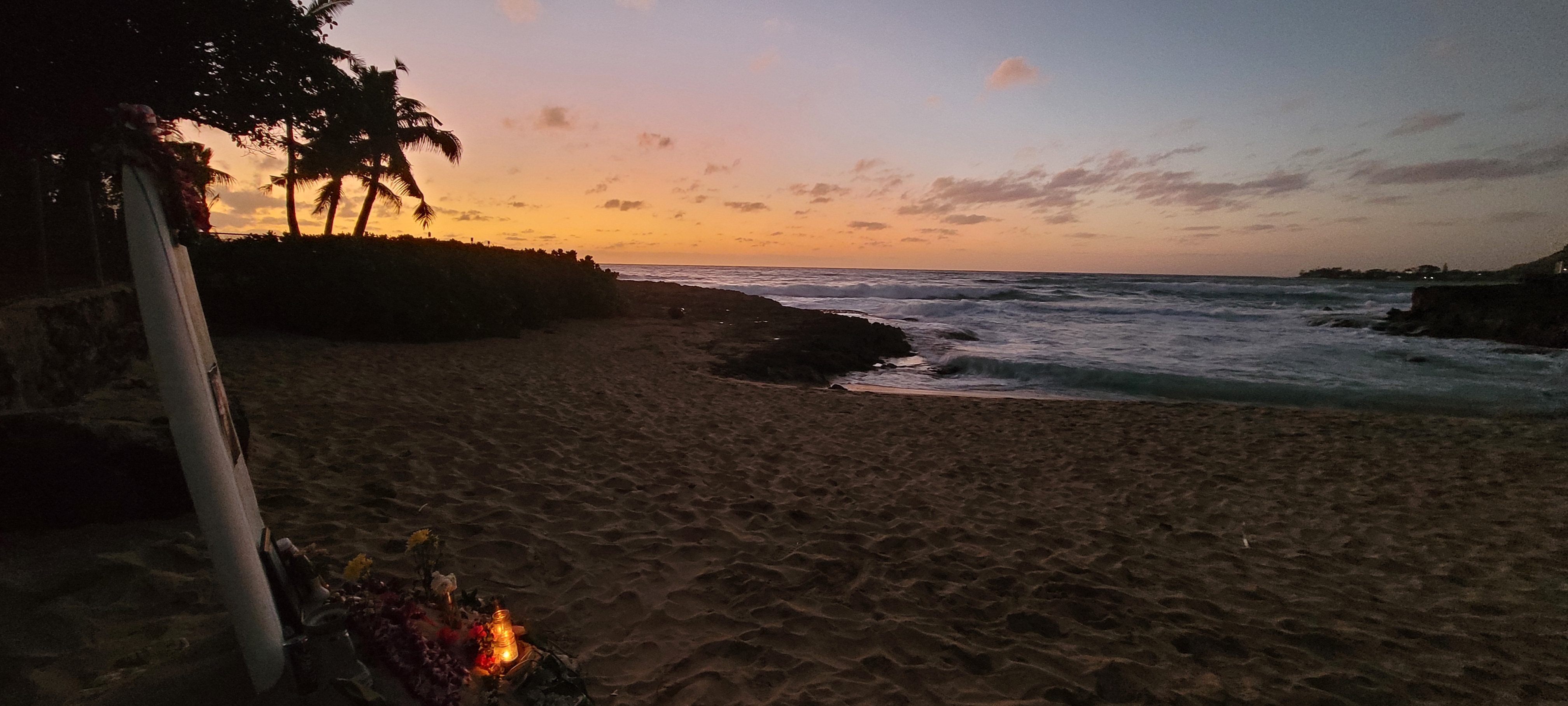 A mound of sand and rocks, with lei and offerings at the base of a surfboard. A wreath is gone now. The sun is setting.
